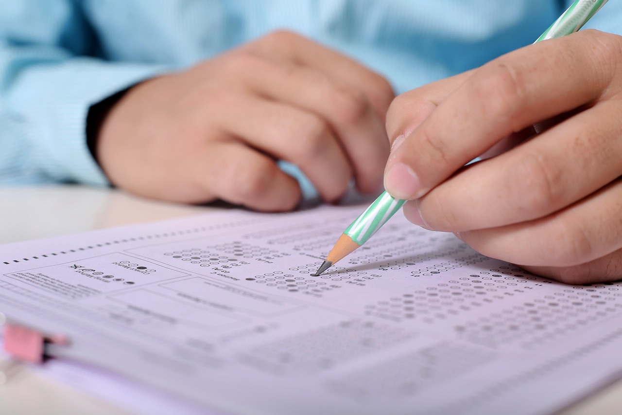 na zdjęciu w tle tablica czarna z napisem EXAM a na pierwszym planie człowiek, w domyśle nauczyciel, stojący z założonymi rękami. rysunek z pixabay.com