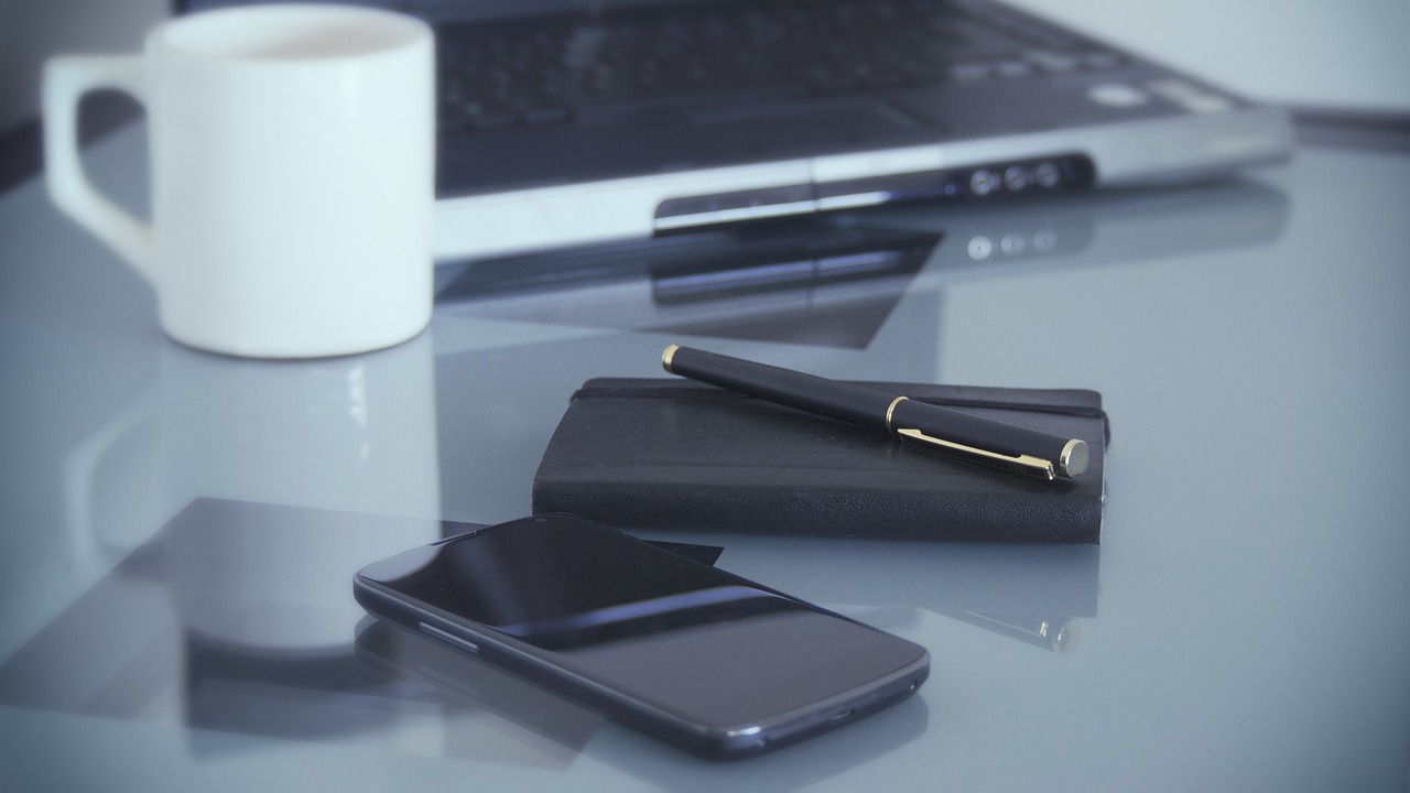 zdjęcie notesu, smarfona, długopisu, kubka na biurku. w tle laptop. zdjęcie z pixabay.com